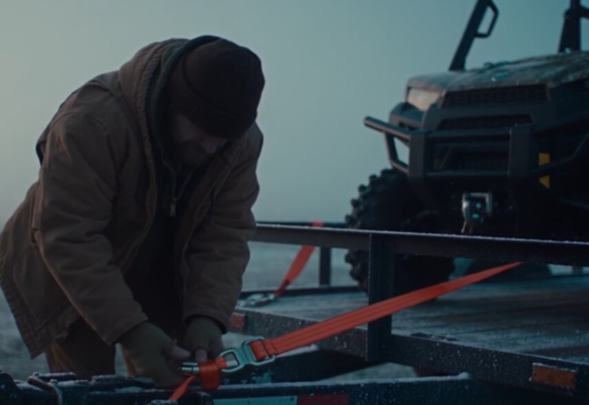 A man in a winter jacket cranking an orange ratchet strap to tie down a side-by-side on a frosty trailer